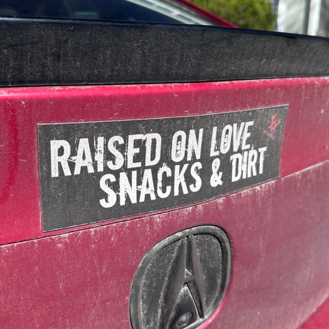Bumper sticker on a car reading 'Raised on Love, Snacks & Dirt' with Acura logo.
