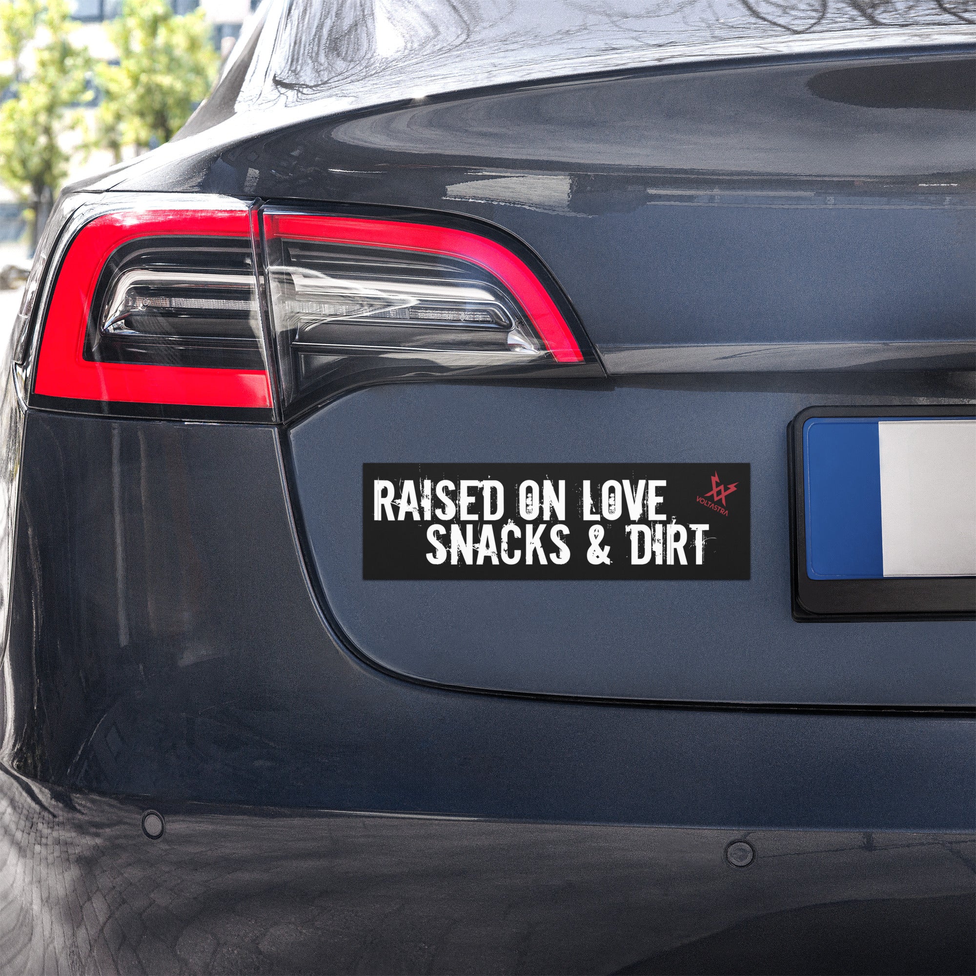 Car rear with a black car magnet that says "Raised on love, snacks & dirt" in a grungy white font and features a red Voltastra logo in the top right corner.