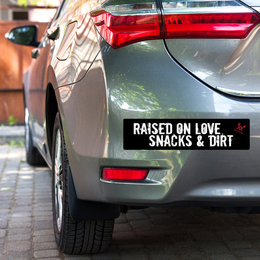 Car with a black car bumper sticker that says "Raised on love, snacks & dirt" in a grungy white font and features a red Voltastra logo in the top right corner.