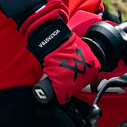 Red gloves with black Voltastra logo gripping a dirt bike throttle