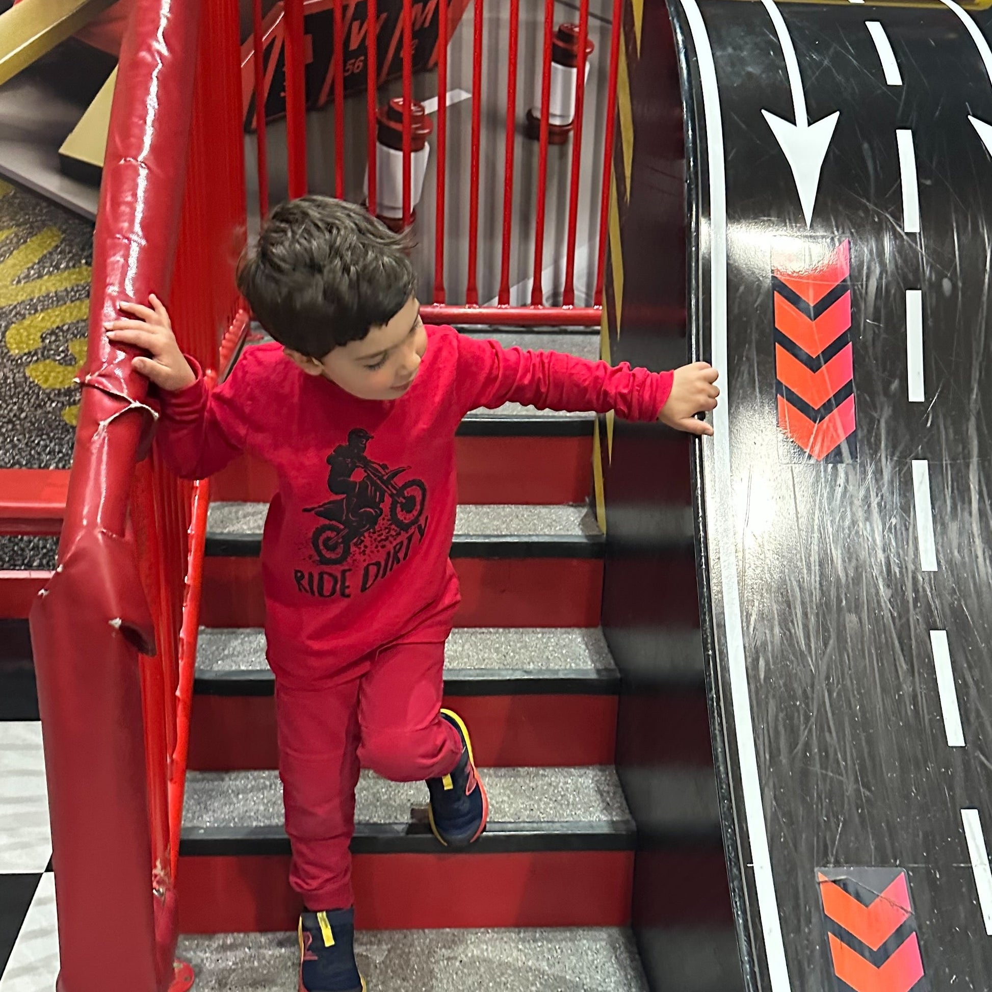 Child playing on a toy race track with red stairs and black and white checkered floor wearing a red t-shirt with a silhouette of a dirt bike riding on top of the words 'Ride Dirty'.